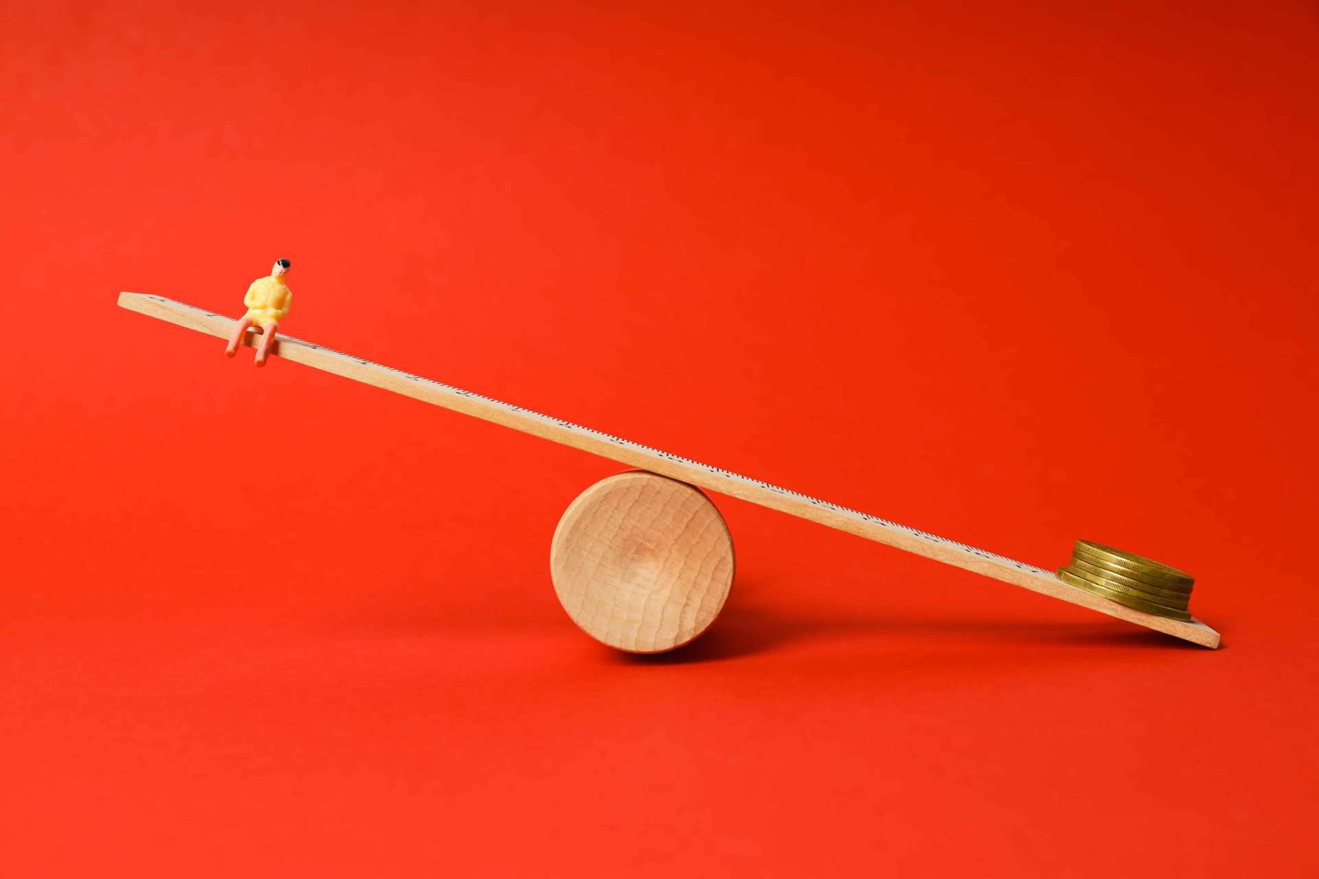 bright red background, timber plank balancing on cylinder. one end is a small person the other is a stack of coins