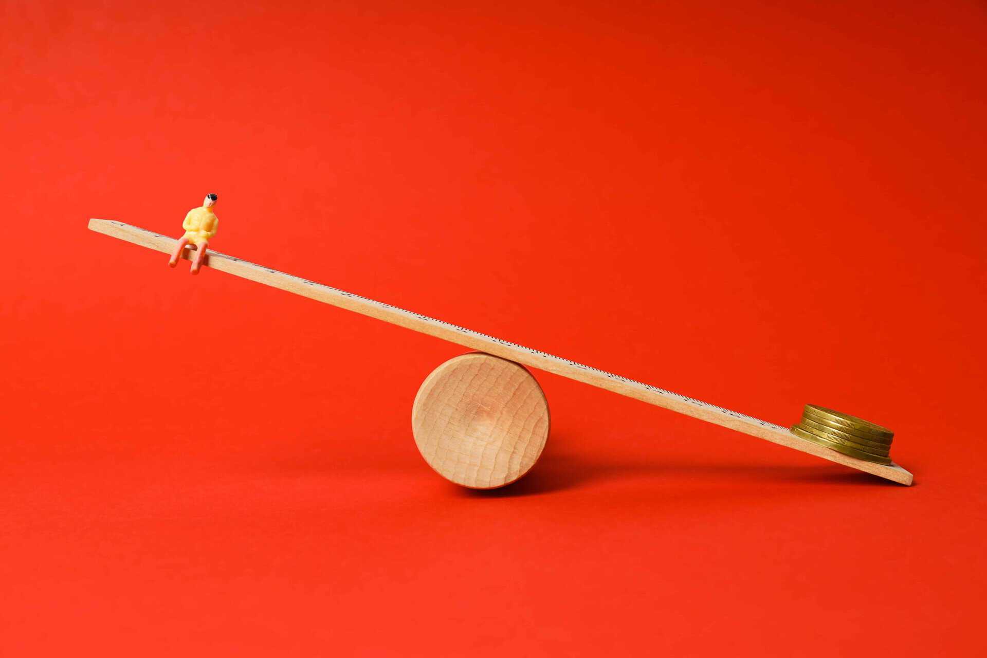 bright red background, timber plank balancing on cylinder. one end is a small person the other is a stack of coins